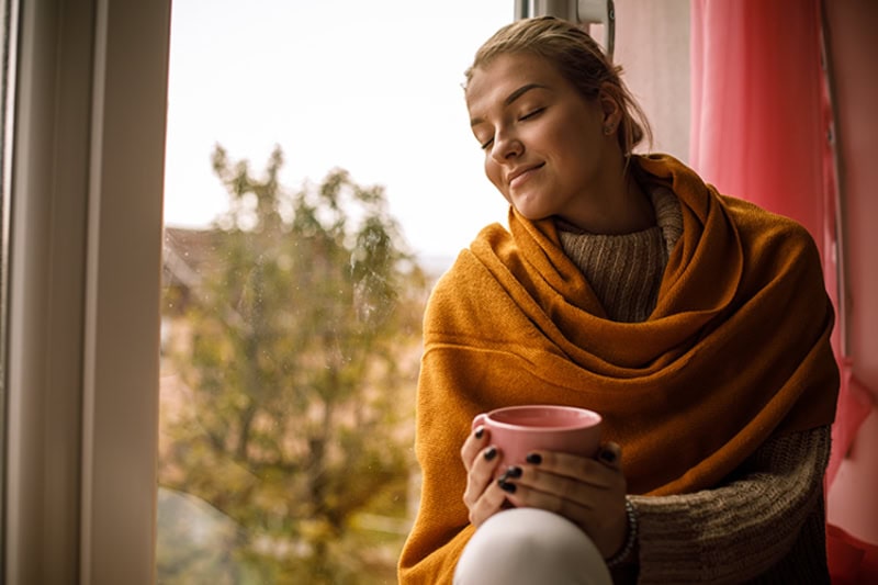 5 Tips to Improve Your Indoor Air Quality This Fall. Photo of a woman sitting on a windowsill looking out a window to a fall view.